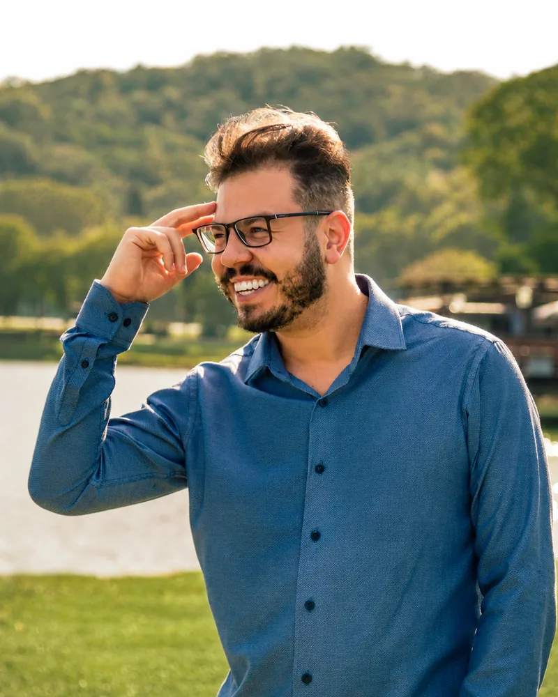 Fabricio Assoni sorrindo ao ar livre, de camisa azul, em um ambiente com árvores e lago ao fundo